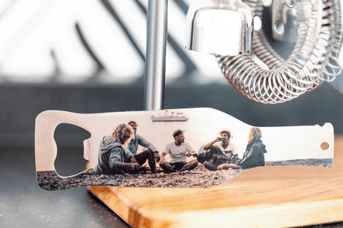 A metal bottle opener sitting on a wooden cutting board. The personalized image on the bottle opener is of a group of friends sitting on a sandy beach talking and laughing. 